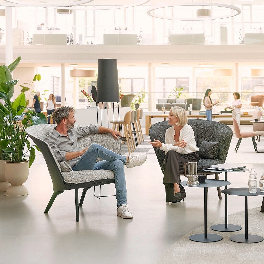 Two people sitting on different upholstered armchairs in a spacious modern office lounge with plants and round tables