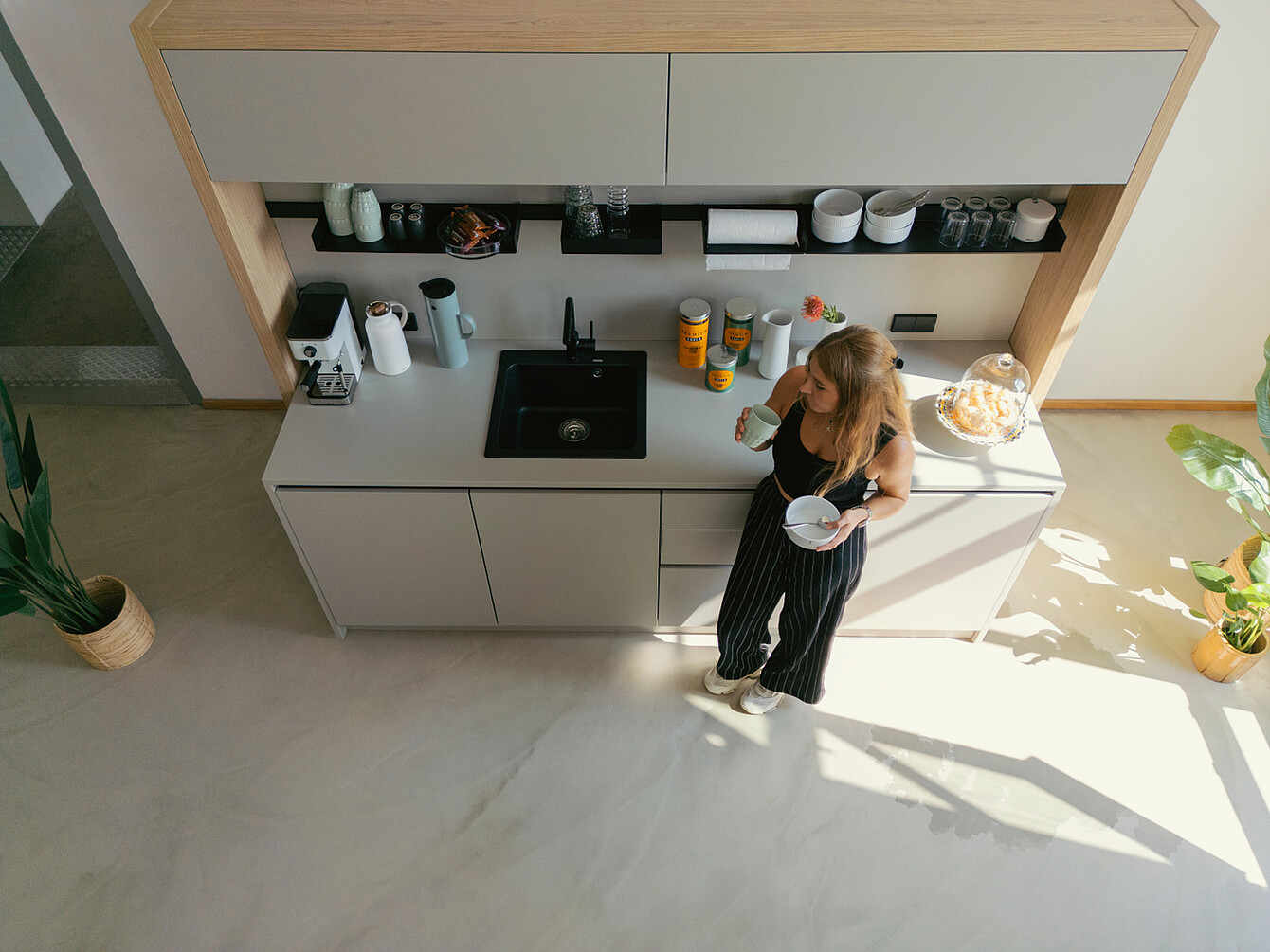 Woman in black top and striped pants standing in a bright office kitchen area with natural light, holding a cup and bowl near a countertop with coffee machine, jars, and plants