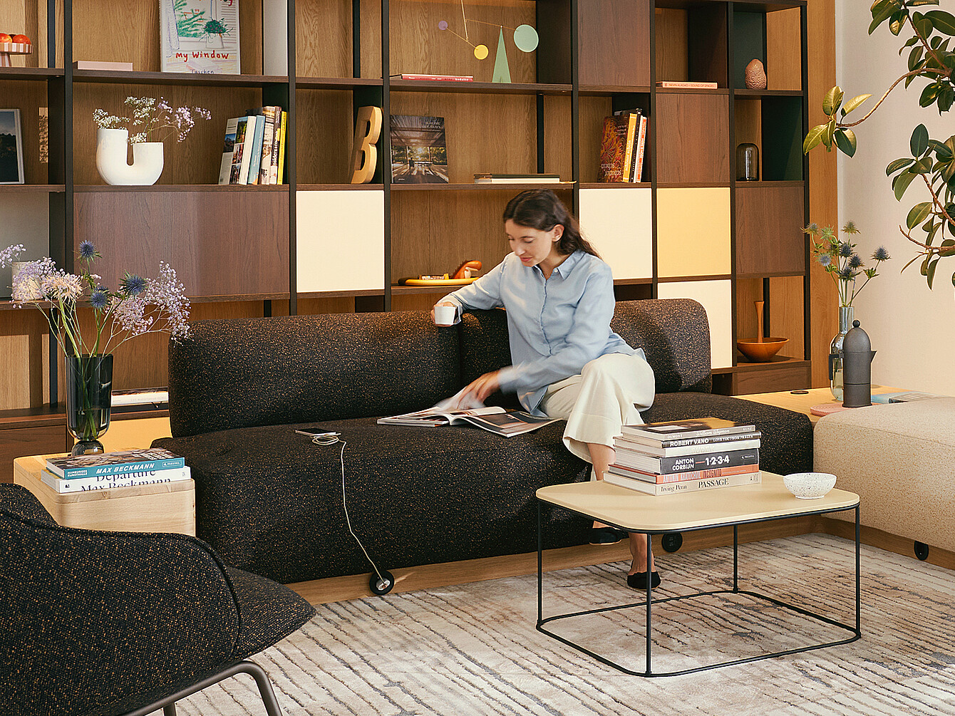 Person sitting on a dark sofa in a work café, reading a magazine with a coffee cup, surrounded by bookshelves and modern furniture