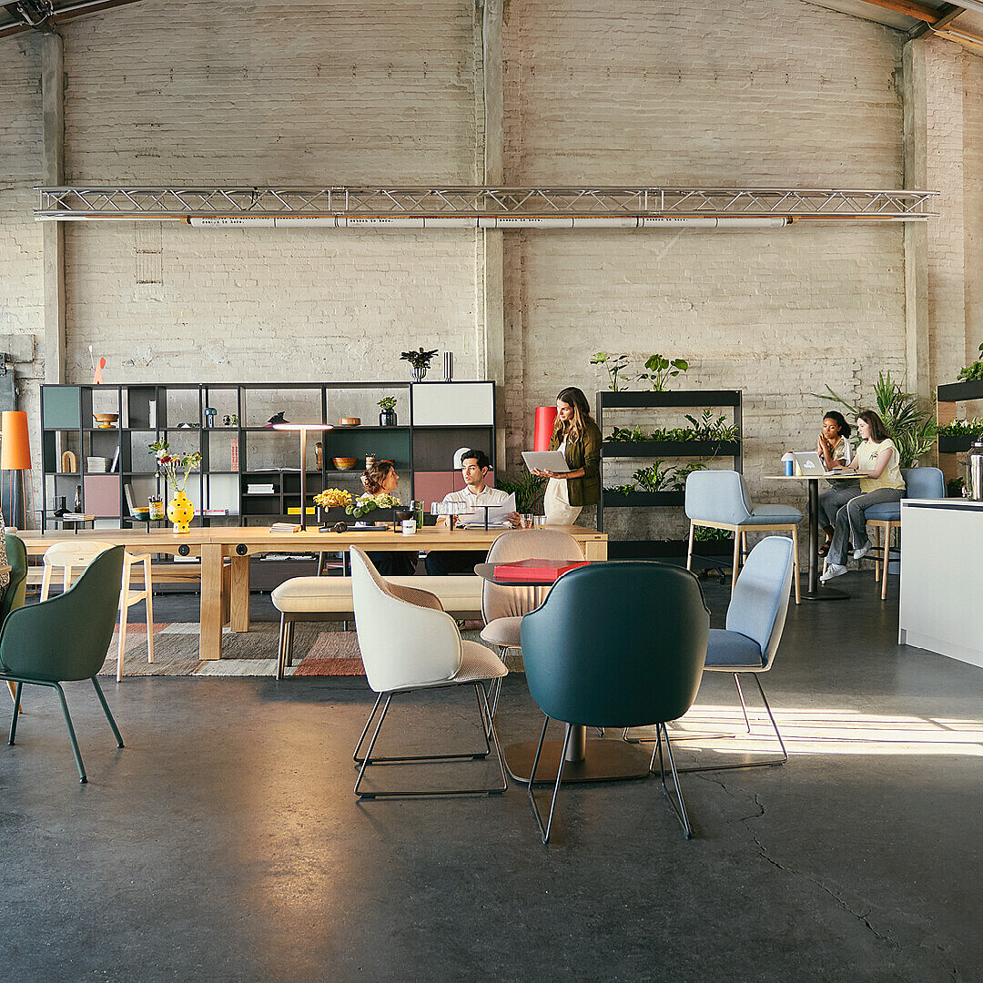 A spacious Work Café featuring various seating arrangements, including colorful chairs and tables. People are engaged in work and conversation, surrounded by plants and modern decor.