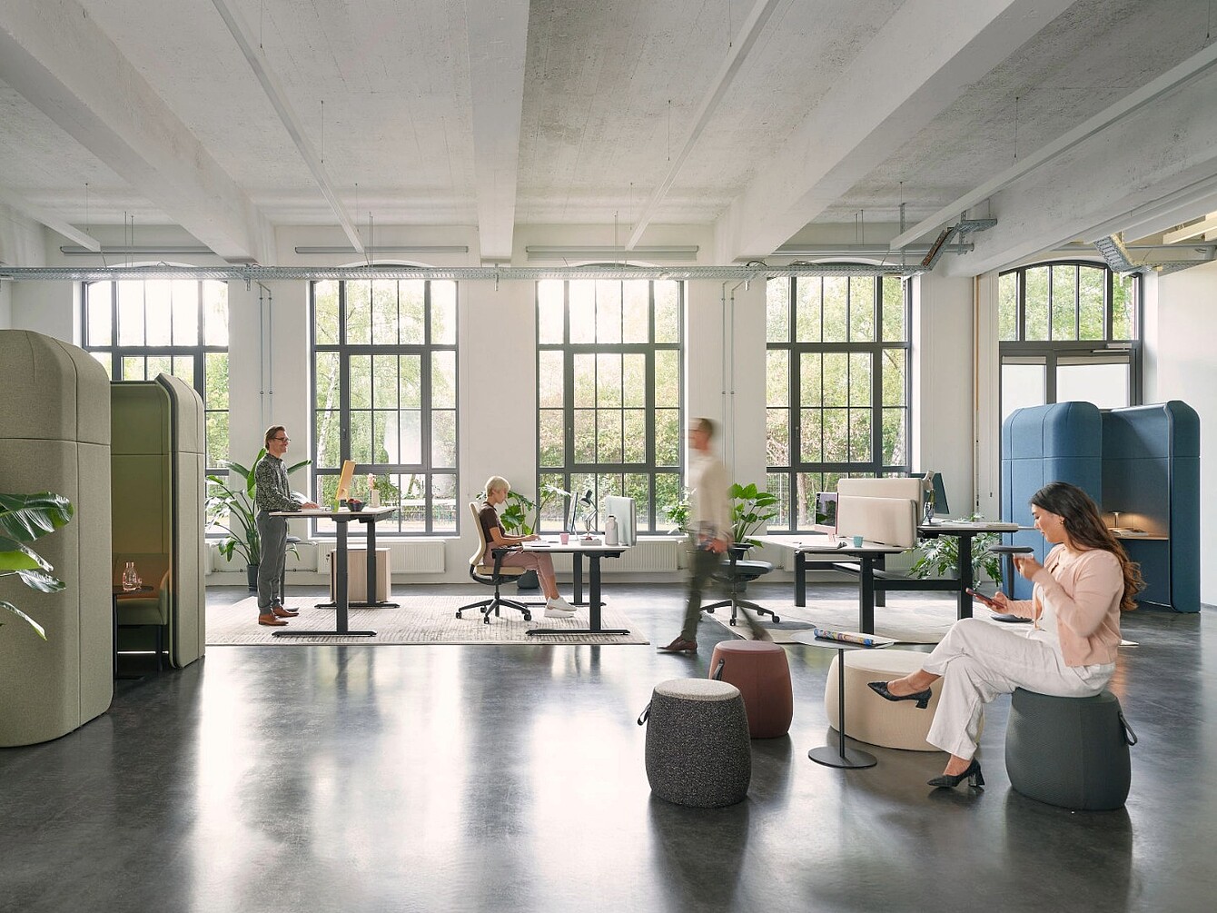Helles Büro mit großen Fenstern, mehreren höhenverstellbaren Schreibtischen, Sitzhockern und einer akustisch abgeschirmten Telefonzelle, Personen arbeiten stehend, sitzend und mit mobilen Geräten