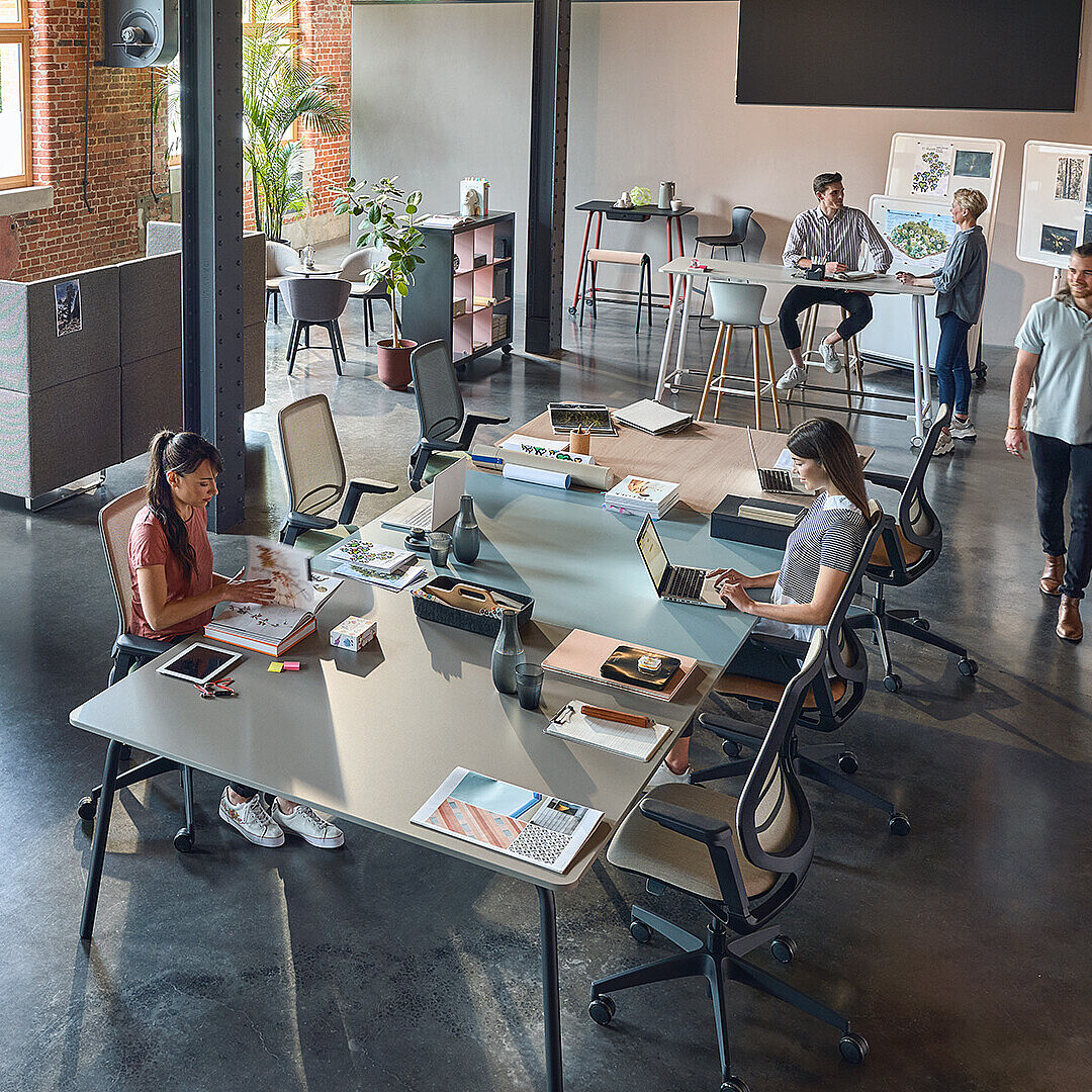 Offenes Büro mit mehreren Personen, die an Computern arbeiten, große Fenster und Backsteinwände, moderne Einrichtung