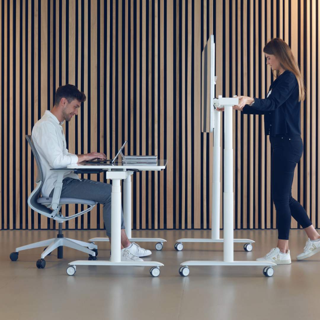 Man sitting at desk working on laptop, woman standing at height-adjustable standing desk in modern office with vertical wooden panels in background