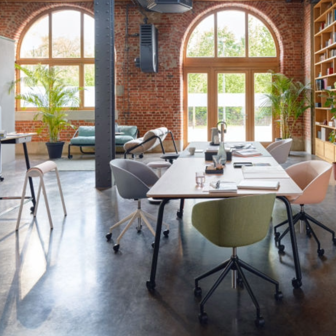 Open-plan office with large arched windows, exposed brick walls, multiple desks with chairs, and plants near the windows