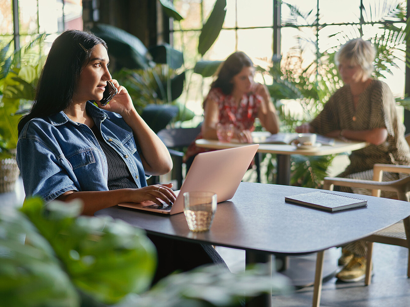 Woman in denim jacket using laptop and talking on phone in a bright cafe with two women working in the background