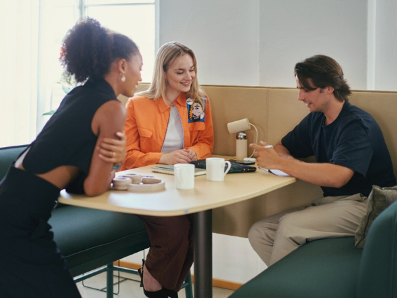Three people sitting at a table in a lounge talking, two women and one man looking at a smartphone.
