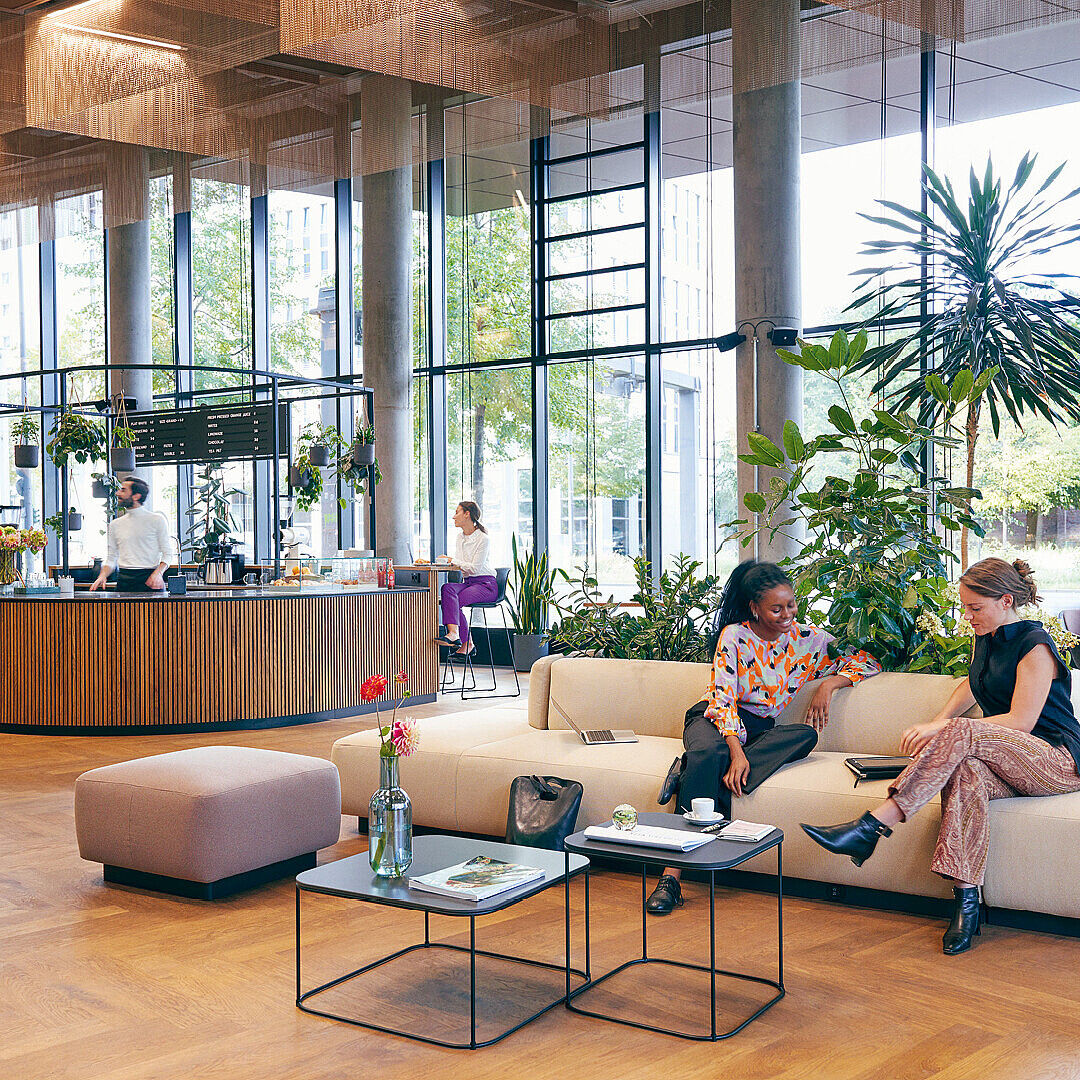 Open modern lounge area with large windows, wooden floor, plants, a curved wooden counter with baristas, and two people sitting on a white sofa talking