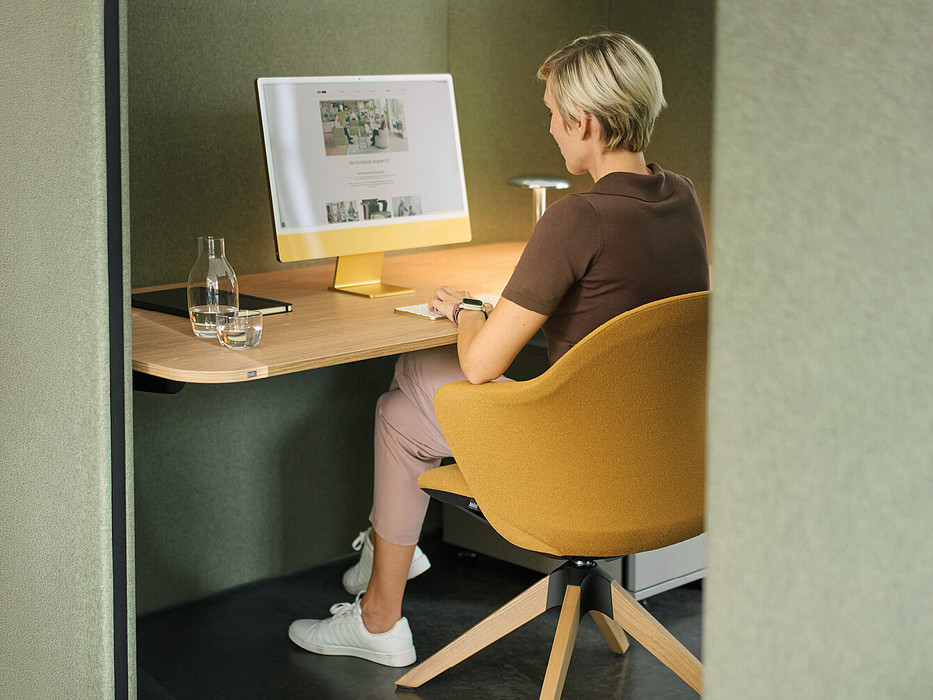 A person with short, blond hair is sitting at a modern desk in a separate, soundproofed workspace. She is wearing a brown T-shirt, beige pants, and white sneakers. The desk chair is yellow and has a rounded shape. On the desk are a large computer with a yellow stand, a glass of water, and a notebook. The walls of the workspace are covered with greenish fabric, creating a calm atmosphere.