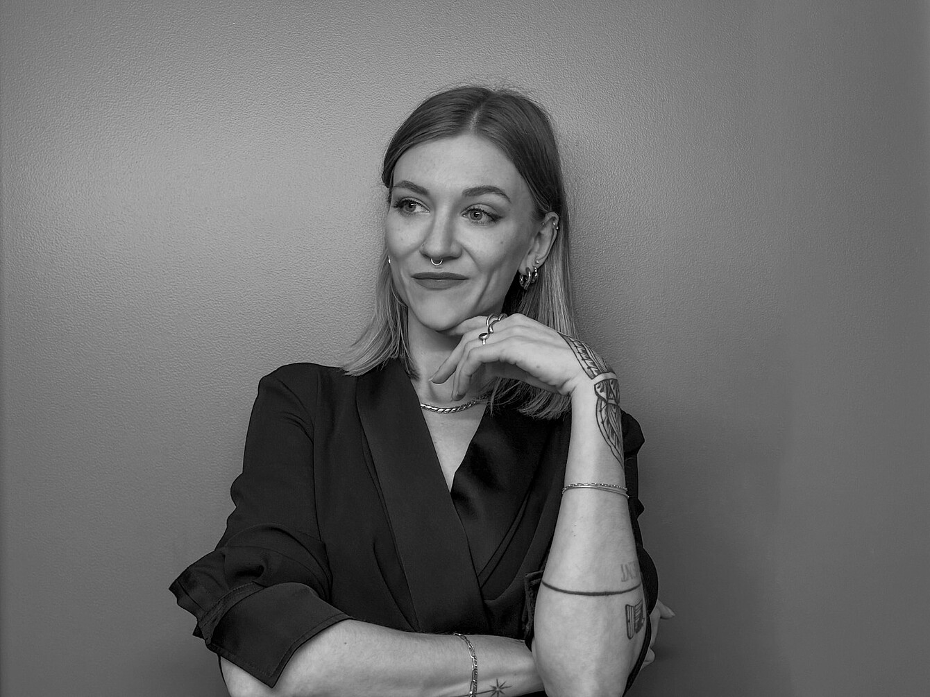 Junge Frau mit langen Haaren, schwarzem Blazer und mehreren Arm- und Handtattoos, die nachdenklich die Hand am Kinn hält.