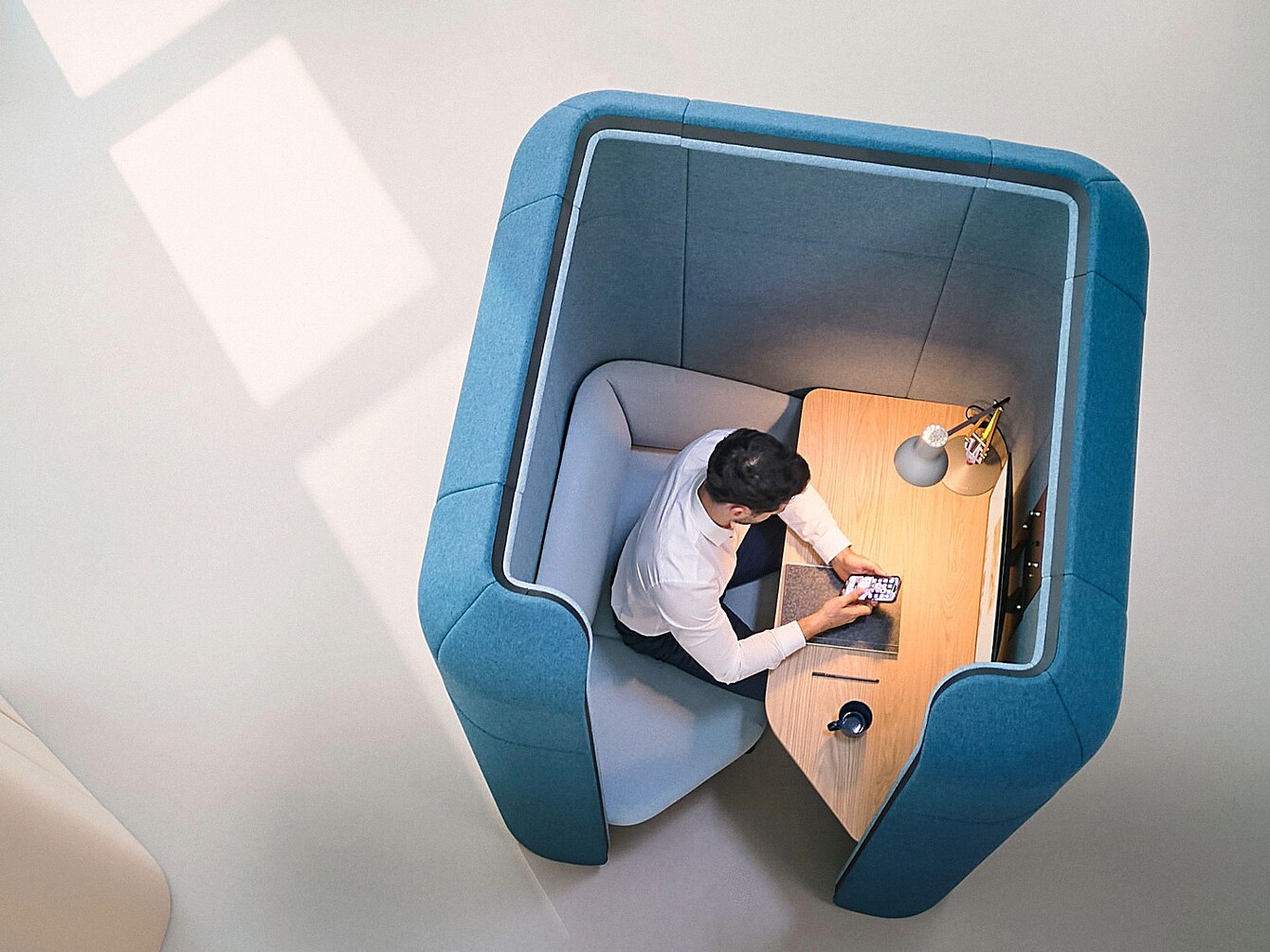 Person sitzt in einem kleinen, abgeschirmten Arbeitsplatz mit gepolsterten blauen Wänden und nutzt ein Smartphone auf einem Holztisch mit Lampe und Tasse