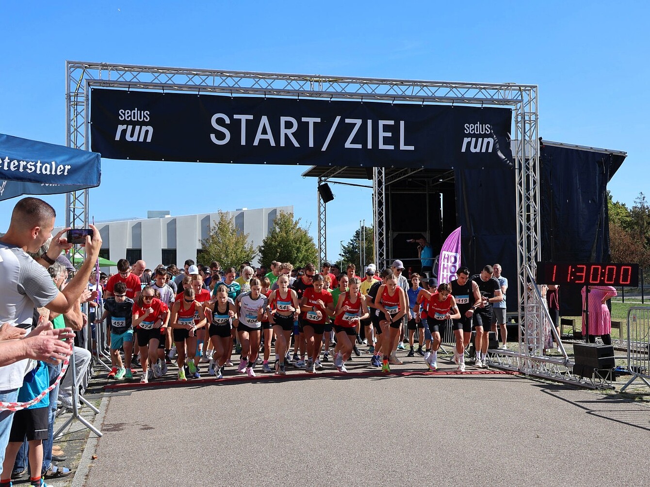 Group of runners at the starting line under a banner reading 'START/ZIEL' with a digital timer showing '1:13:00'
