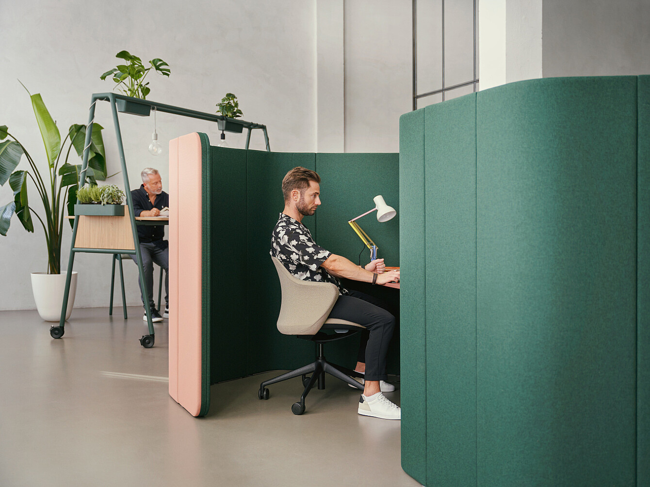 Man working in a green acoustic sanctuary pod in a modern office with natural light and plants, reflecting workplace wellbeing priorities.