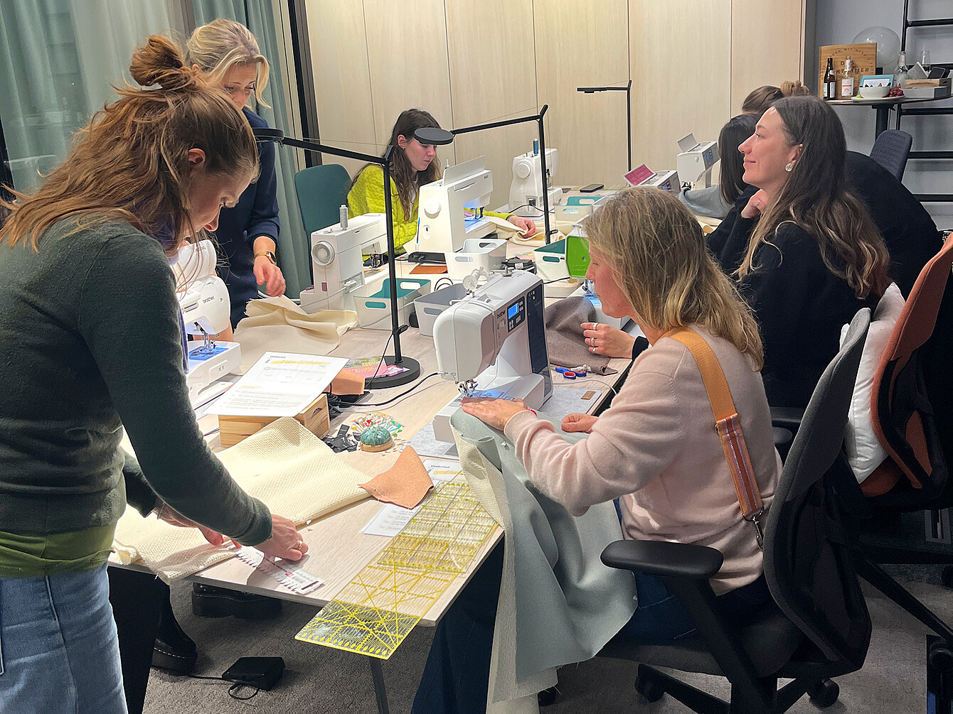 Mehrere Personen sitzen an einem Tisch mit Nähmaschinen, Stoffen und Schneidwerkzeugen in einem Innenraum, sie arbeiten gemeinsam an Textilprojekten.