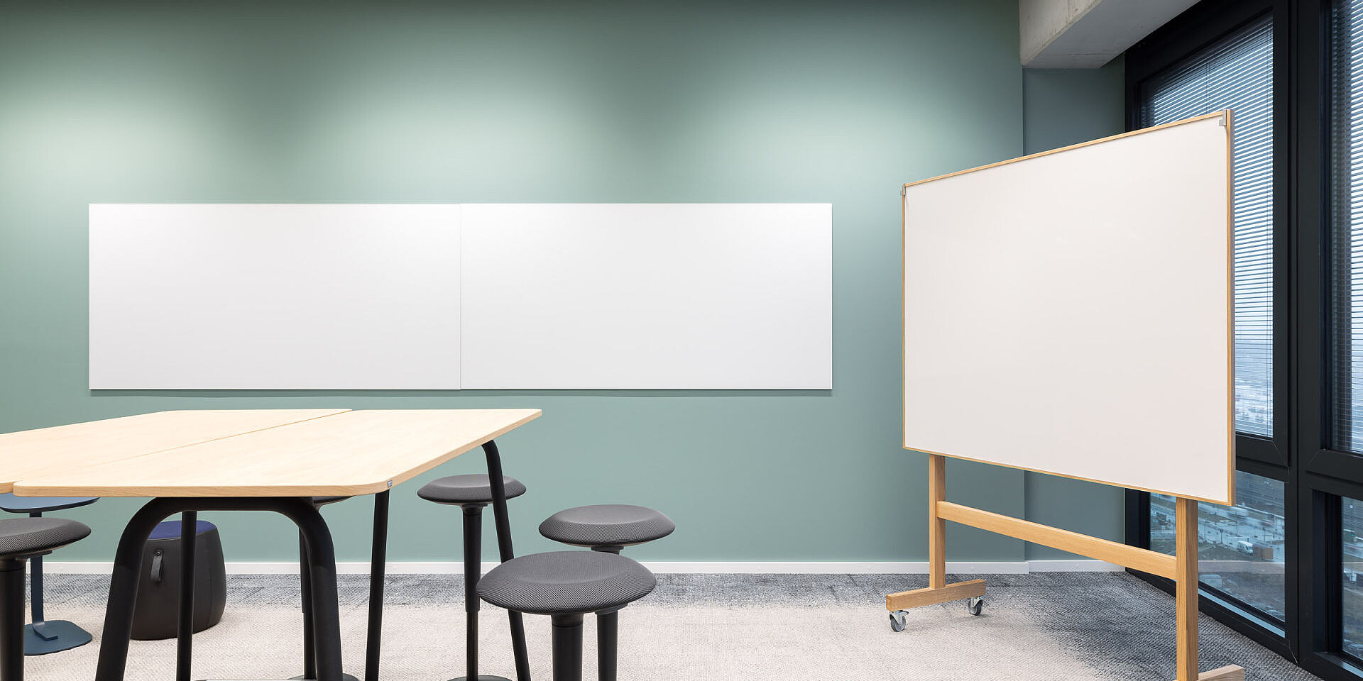 Moderner Arbeitsbereich mit se:lab high desk Workshop-Tisch, se:fit Stehsitz und mehreren universellen se:dot Hockern vor grüner Wand und mobilem Whiteboard 