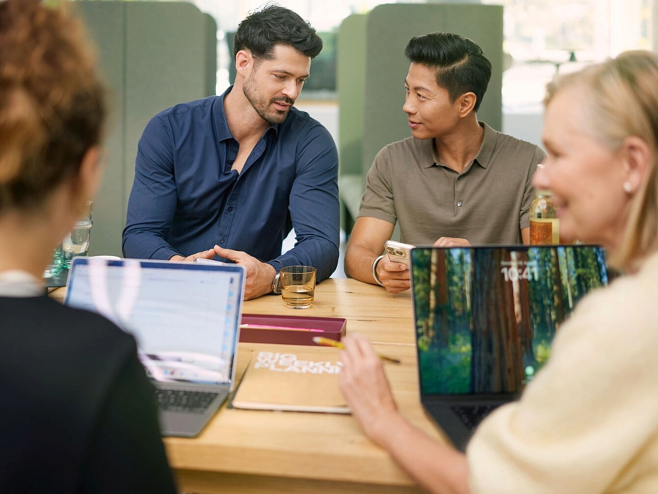 Four people sitting around a wooden table in an office with laptops and notebooks, two facing the camera and two with backs turned