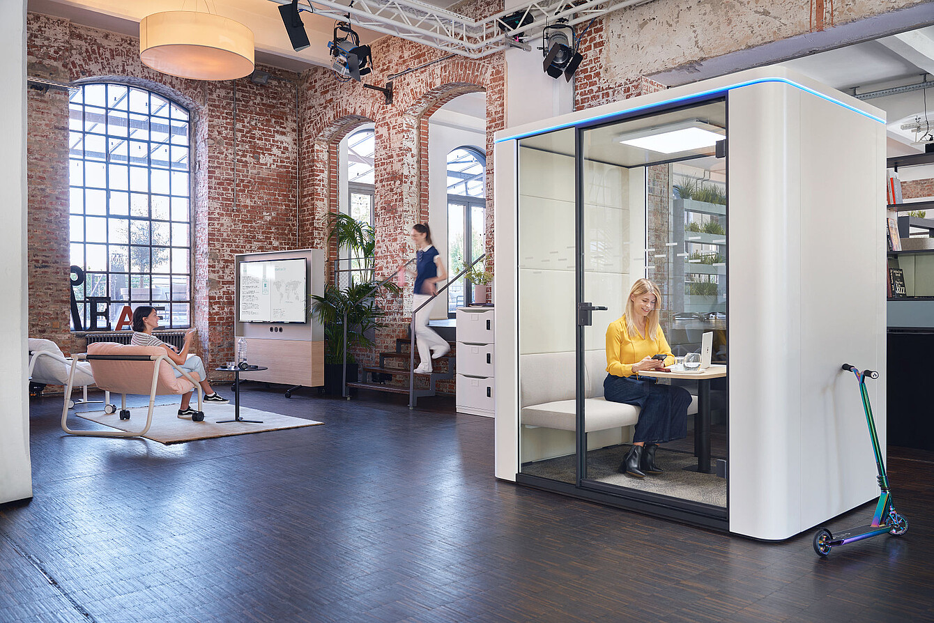 Moderne Bürofläche mit Backsteinwänden, zwei Personen in se:cube Raum-in-Raum-Lösungen, eine Frau arbeitet am Laptop in einem abgetrennten Raum mit Touchdisplay zur Steuerung von Beleuchtung und Belüftung, ein E-Scooter steht daneben
