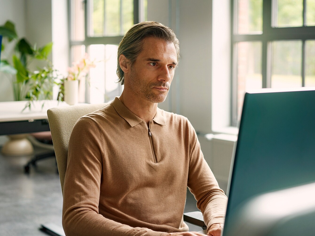 Middle-aged man wearing a brown polo shirt sitting focused in front of a computer in a bright office with large windows.