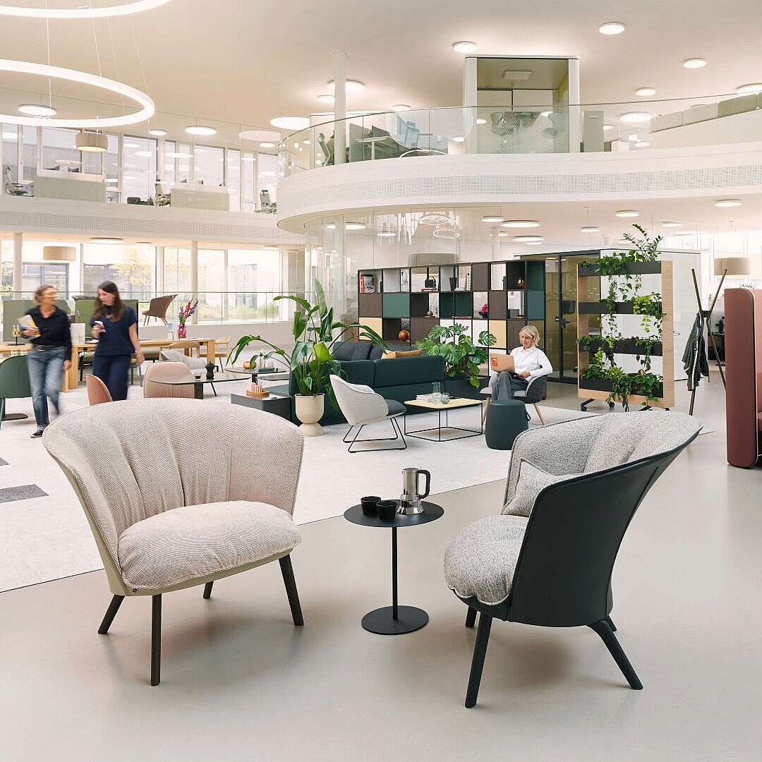 Offener moderner Bürobereich mit mehreren Sitzgruppen, darunter zwei gepolsterte Sessel im Vordergrund und eine halbhohe, mit Stoff verkleidete Co-Creation-Nische mit zwei Personen am Tisch