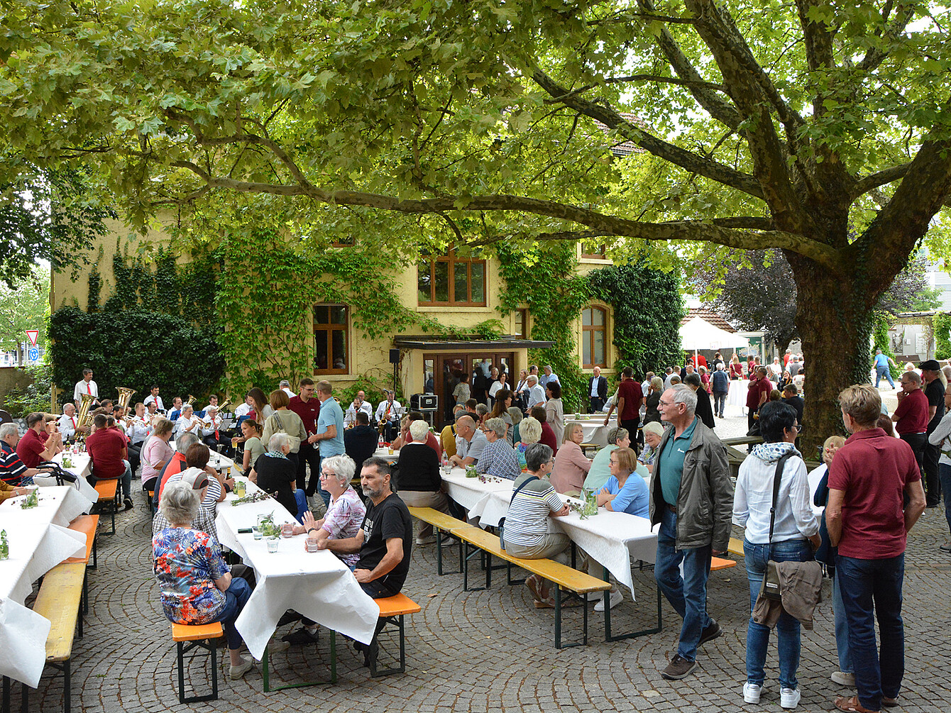 Gäste sitzen und stehen bei Sedus Werksmusik Jubiläumskonzert im Garten mit Tischen, weißen Tischdecken und Bäumen, genießen Getränke und Speisen