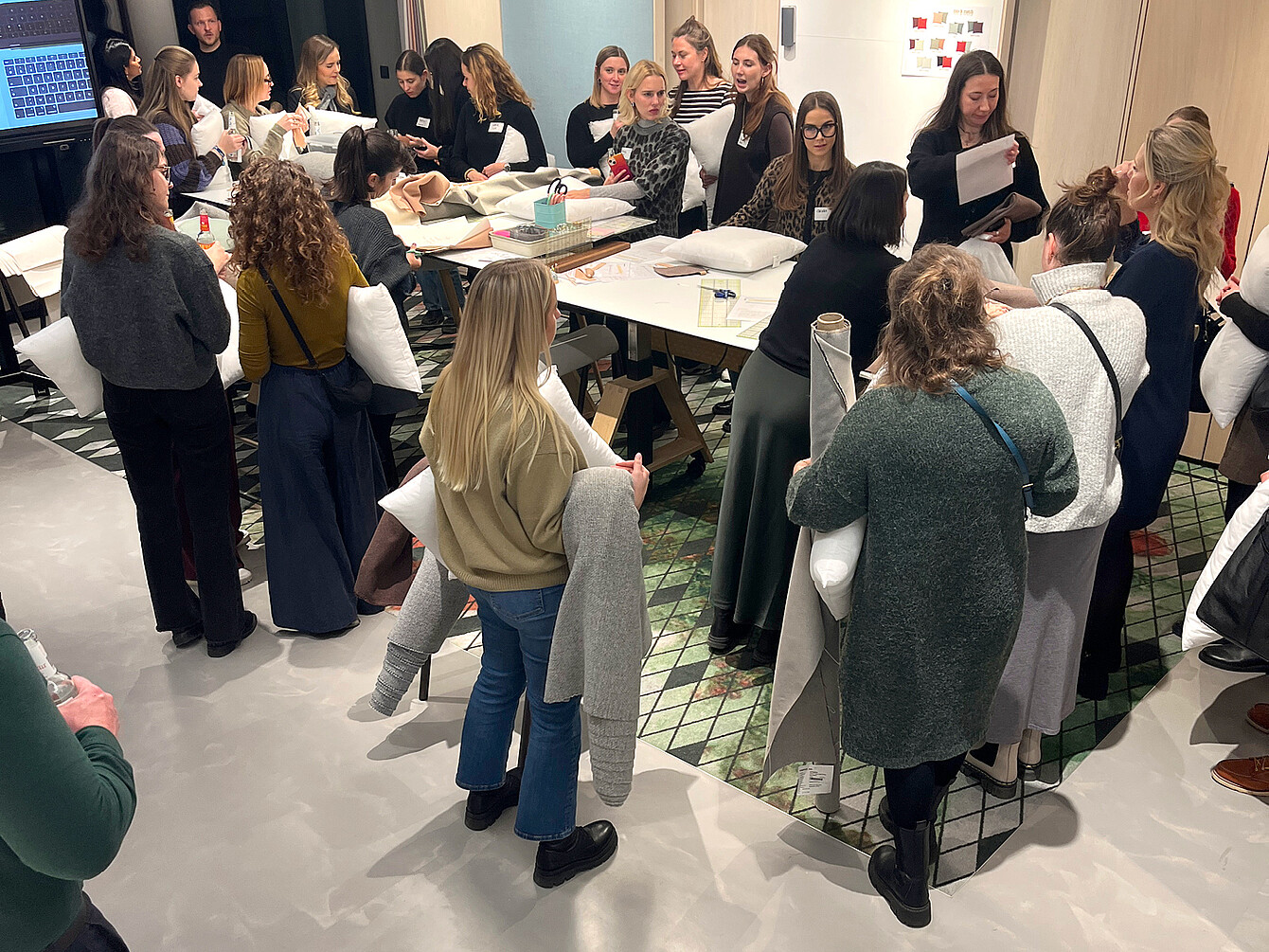 Mehrere Personen stehen und sitzen um einen großen Tisch mit Kissen und Papieren, in einem Raum mit gemustertem Teppich und hellen Wänden, bei einem Event von Sedus und Gabriel in München.