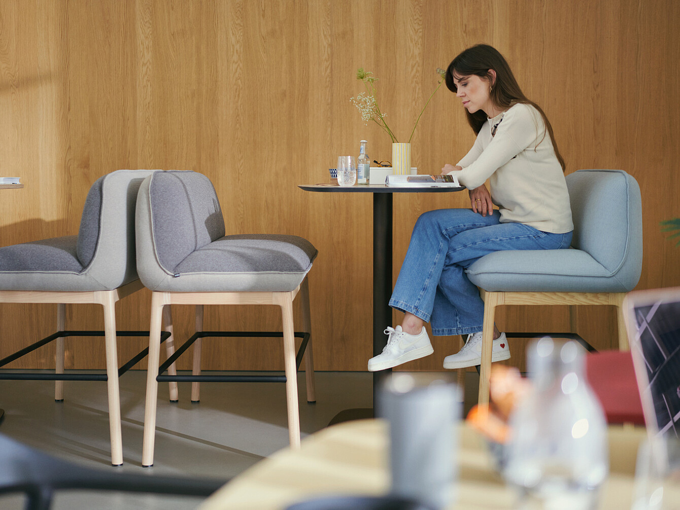 Woman sitting alone at a small table in a modern workplace lounge with wooden walls, reading or working quietly, illustrating a calm and supportive environment for employee wellbeing.