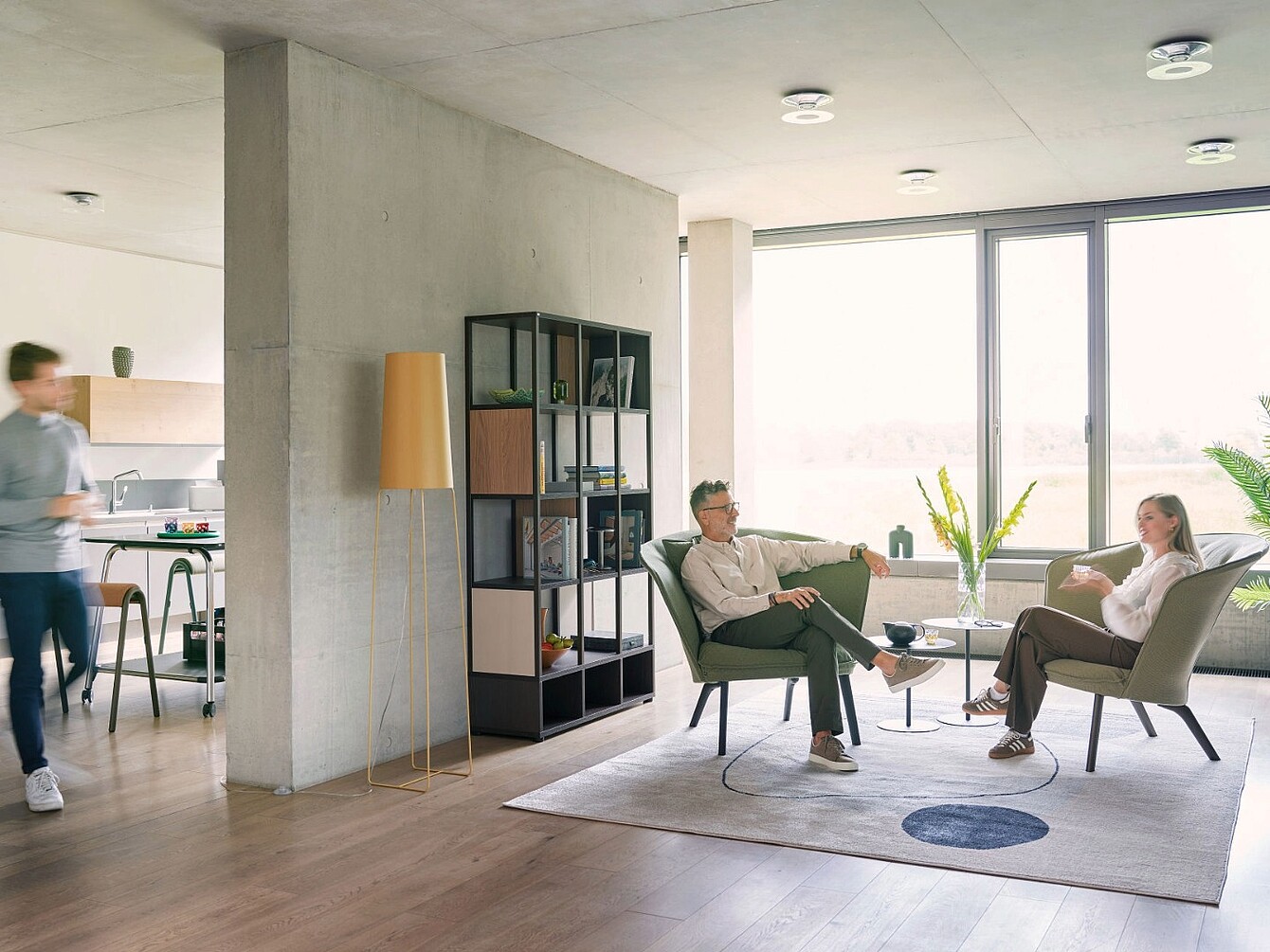 Two people sitting in modern armchairs facing each other in a bright room with large windows, a rug, a floor lamp, and a shelving unit, with a blurred person walking in the background near a kitchen area