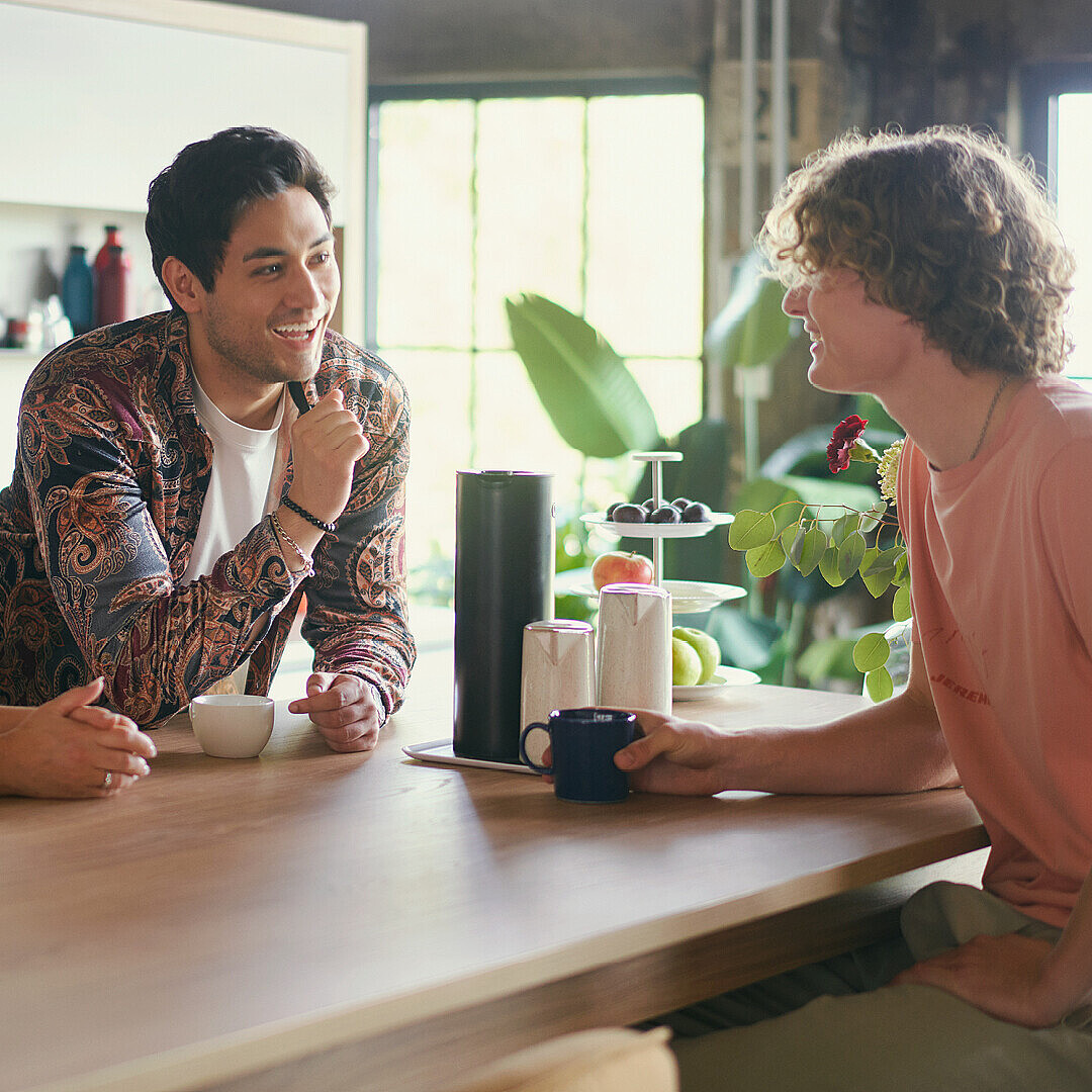 Drei Personen sitzen an einem Holztisch in einem hellen Büro mit großen Fenstern, trinken Kaffee und unterhalten sich in entspannter Atmosphäre