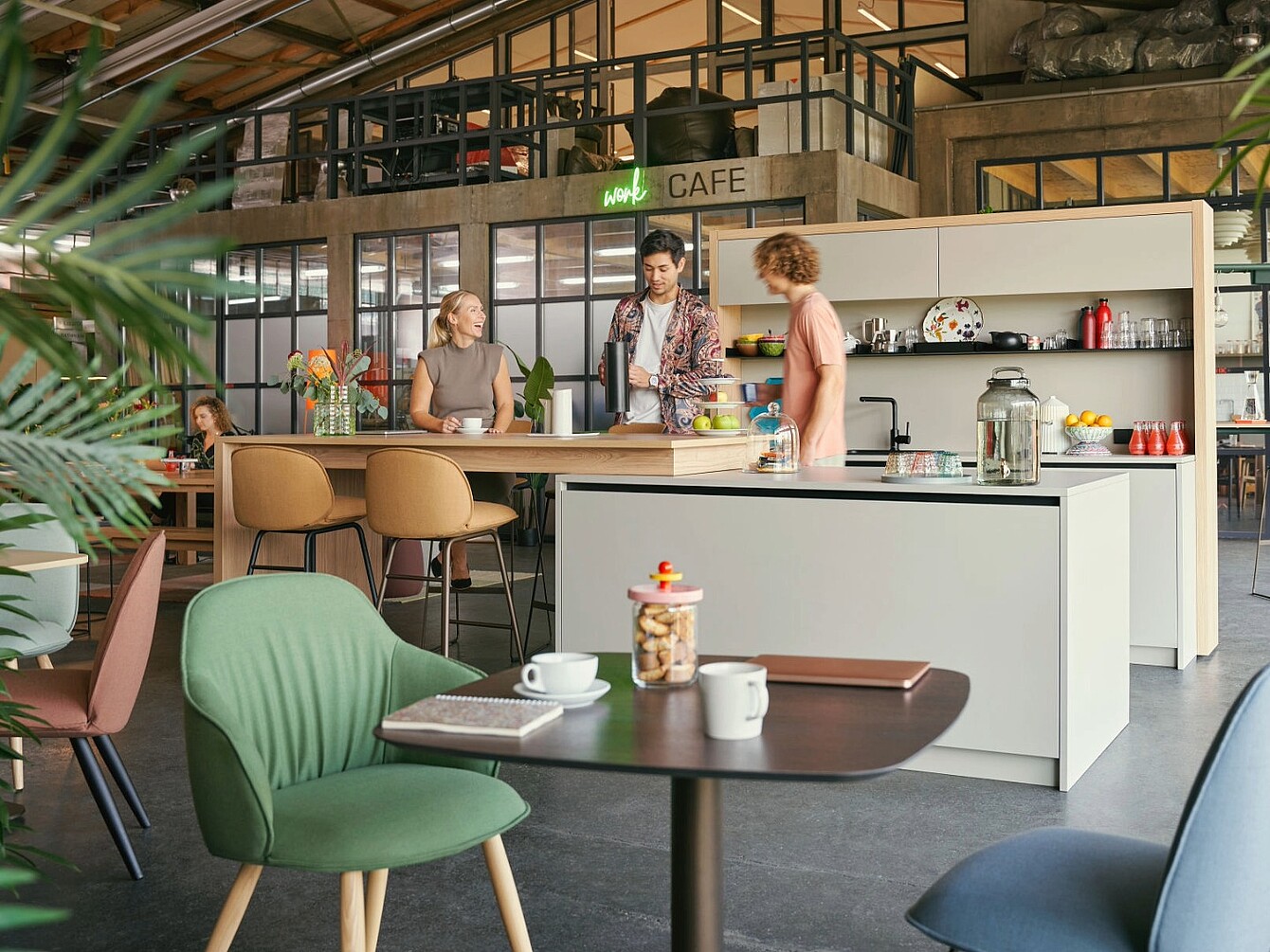 Drei Personen stehen an einer modernen Kücheninsel in einem offenen Büro mit Café-Schild und sitzen zwei weitere Personen an Tischen mit Tassen und Büchern
