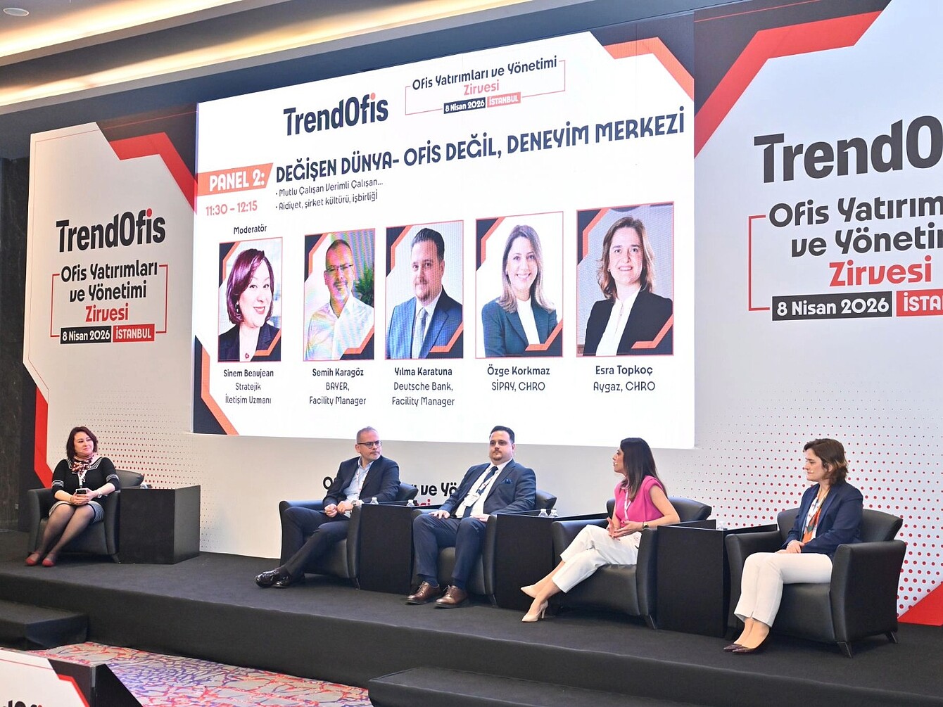 Fünf Personen sitzen auf einer Bühne vor einer großen Wand mit dem Schriftzug 'TrendOfis' und weiteren Texten zum Thema Büroinvestitionen und -management, vier von ihnen auf Sesseln, eine auf einem Hocker, alle in formeller Kleidung, im Hintergrund sind Porträtfotos und Namen der Panelteilnehmer zu sehen.