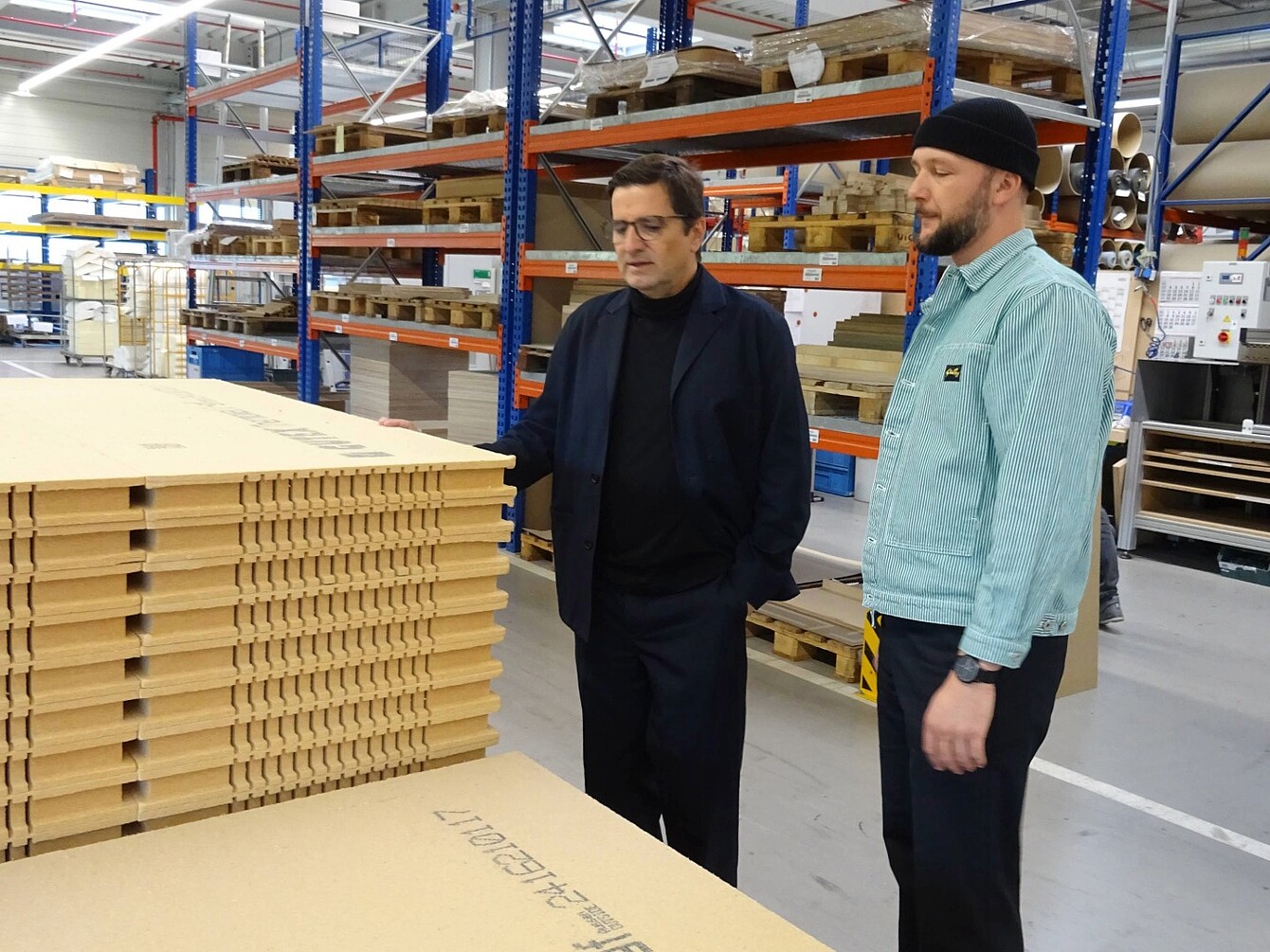 Zwei Männer (Rudi Raschke und Konstantin Thomas) stehen in einer Lagerhalle vor einem Stapel Holzfaserplatten, umgeben von Regalen mit weiteren Holzmaterialien.
