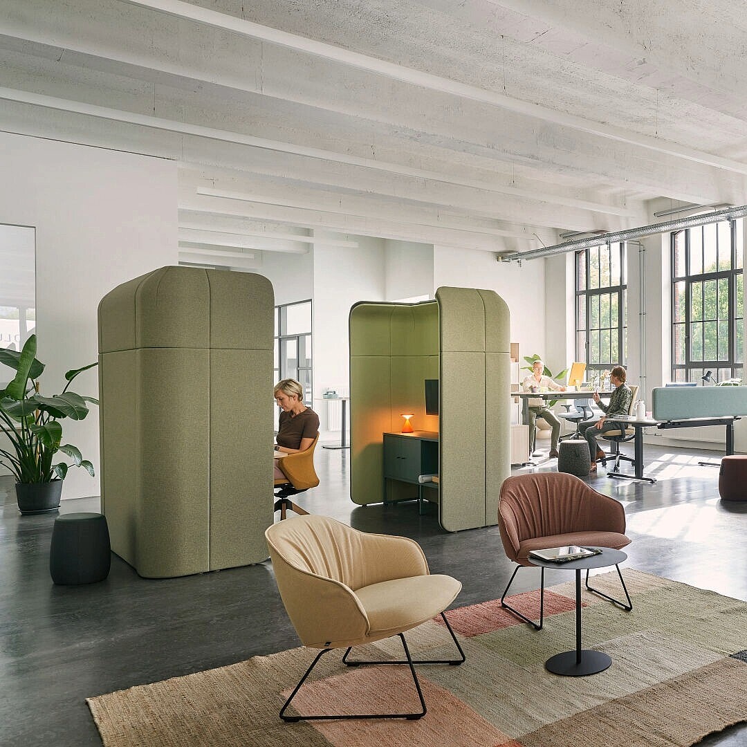 Open-plan office with large windows, featuring textile privacy pods in green and blue, and seating areas with modern chairs and small tables on a rug.