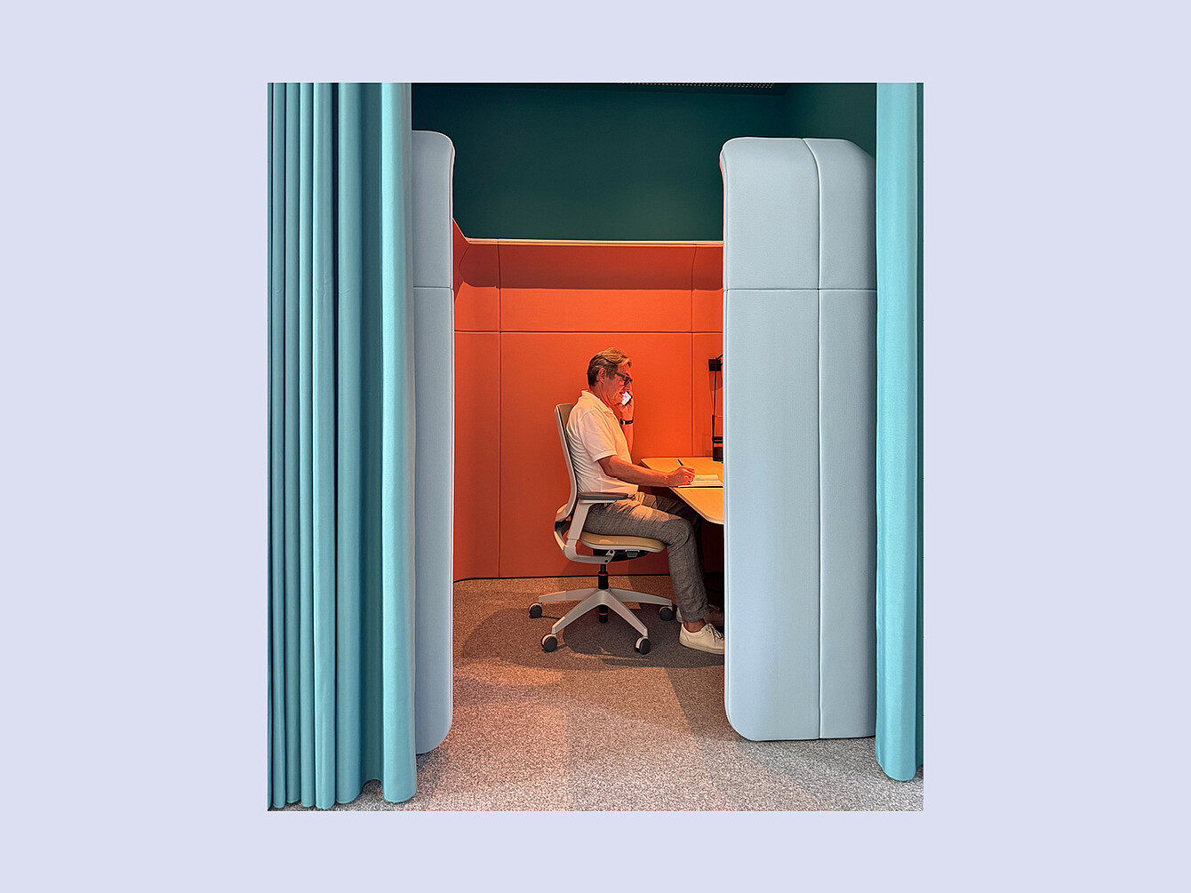 Person sitzt an einem Schreibtisch in einer modernen, schallisolierten Arbeitskabine mit oranger Rückwand und hellblauen Seitenwänden