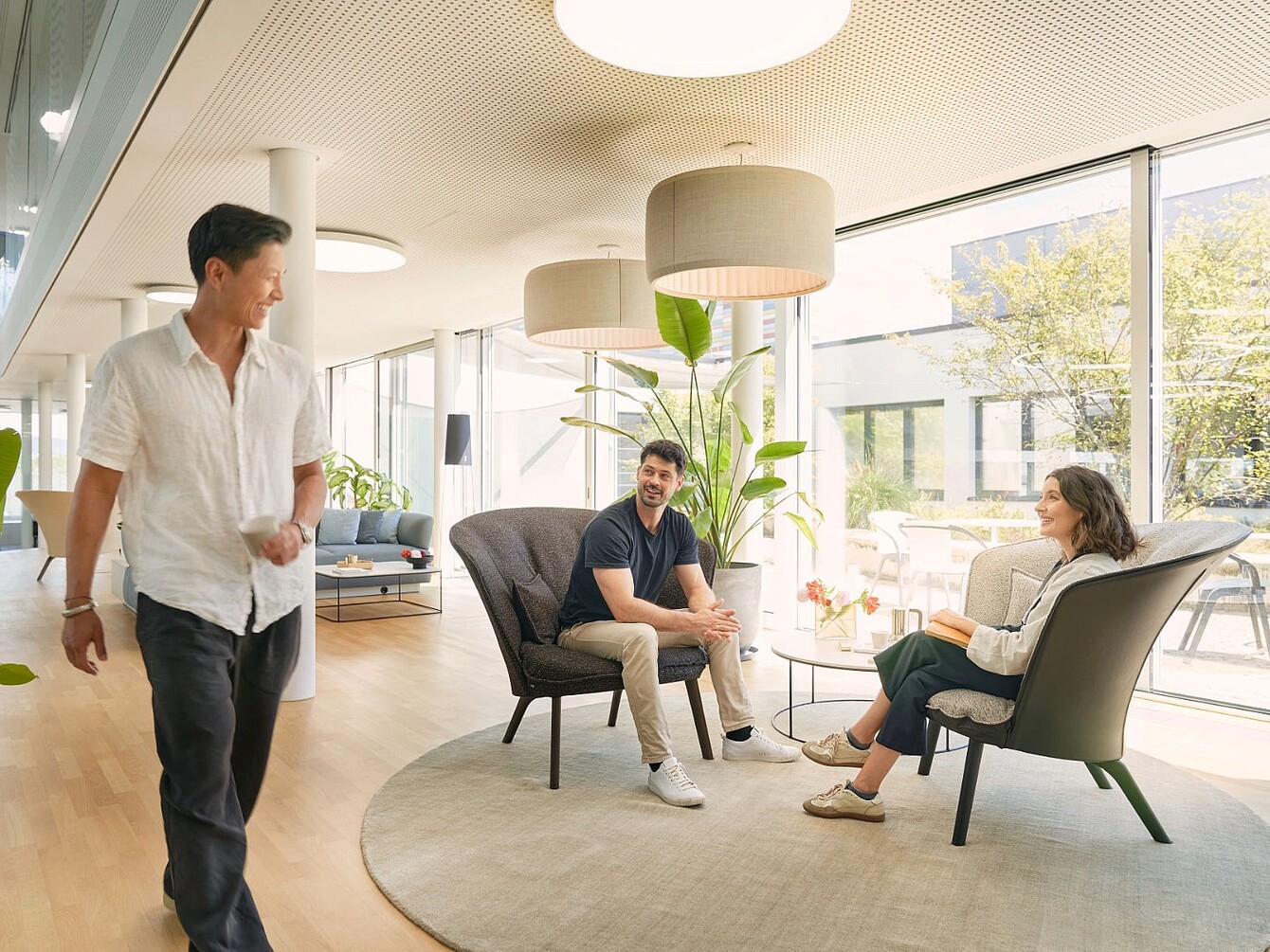 Three people in a modern office lounge with large windows sitting and standing around a round rug and armchairs