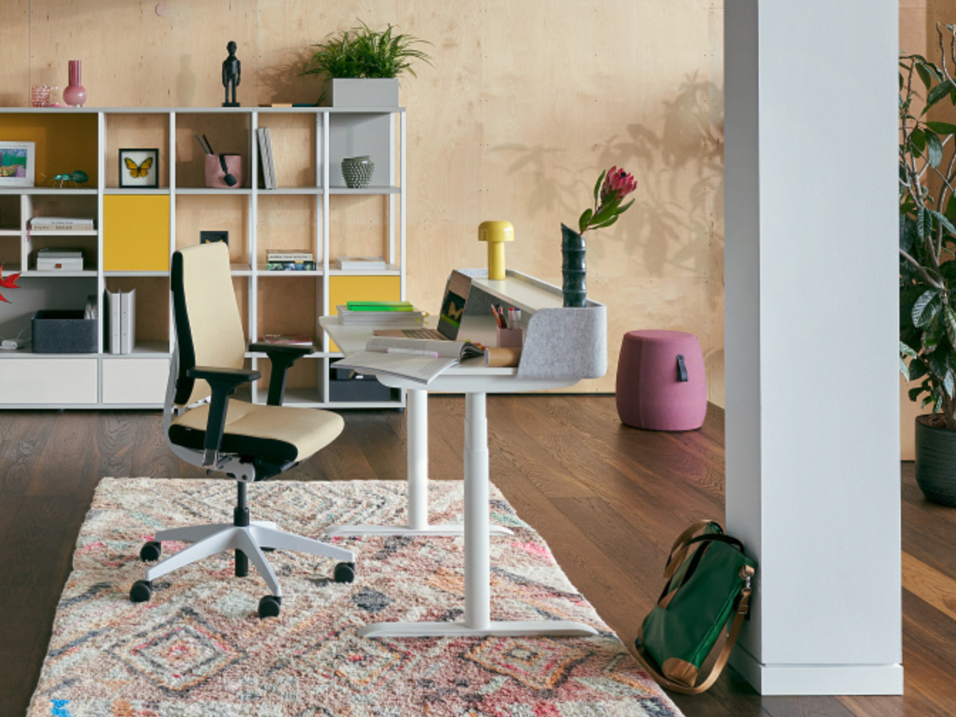Modern workspace with height-adjustable desk, ergonomic office chair, geometric-patterned rug, and shelving unit with colorful compartments in the background