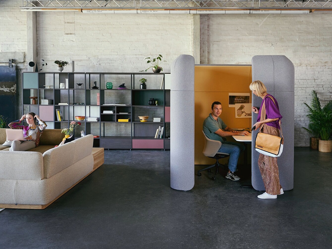 Zwei Personen in einem modernen Büro, eine sitzt in einem halb geschlossenen, schallabsorbierenden Arbeitsplatz, die andere steht davor und spricht mit ihr, im Hintergrund ein Sofa und ein großes Regal mit Pflanzen und Dekoration.