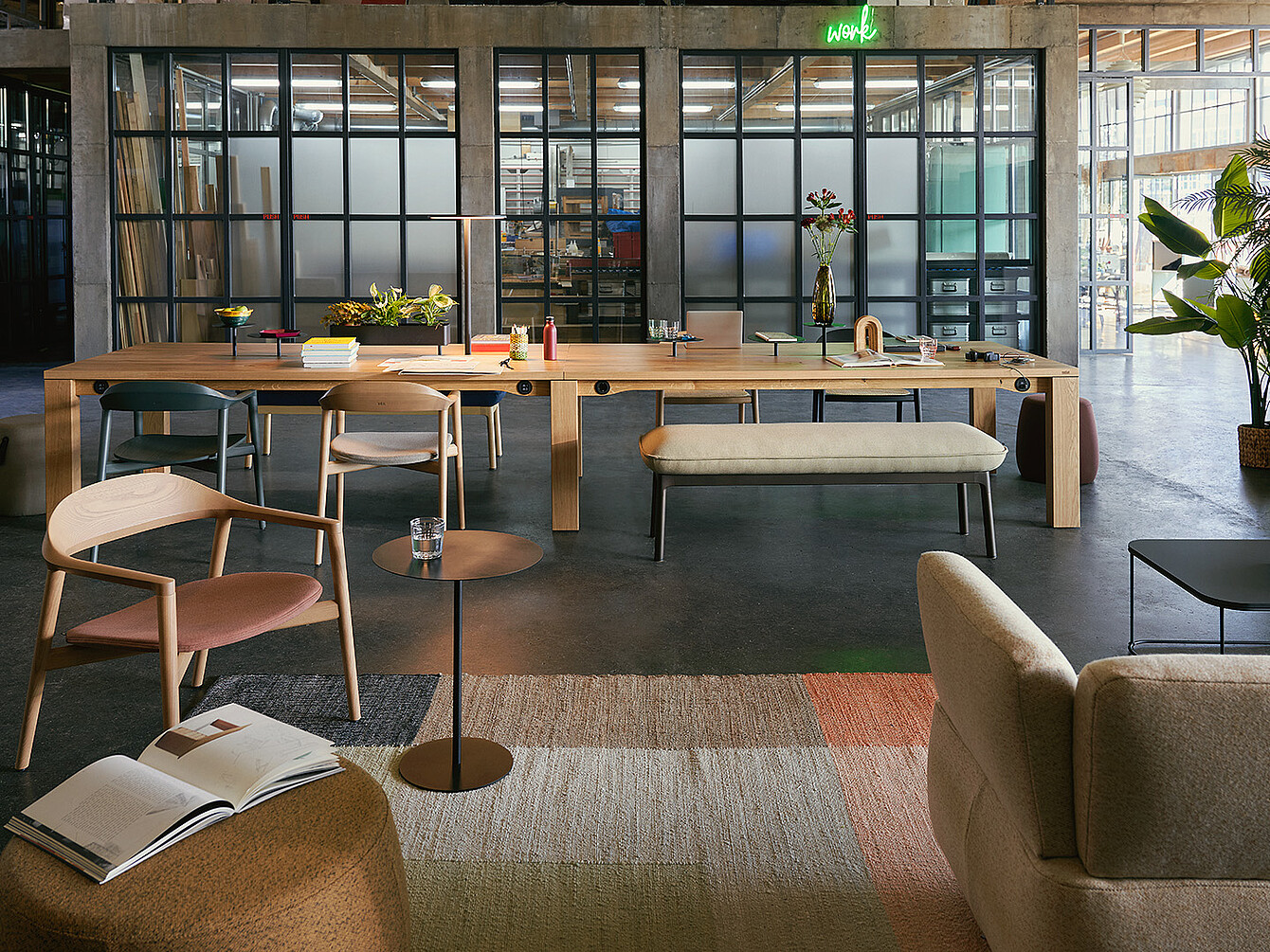 Modern office lounge area with wooden chairs, a long wooden table with books and flowers, and a glass wall with black frames