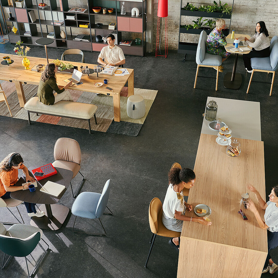 Open office space with people working and talking at tables and a kitchen area with coffee machine and fruit bowl