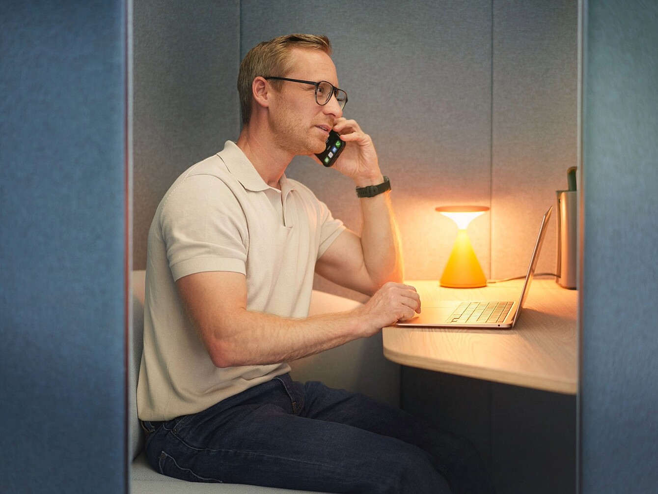 Man wearing glasses sitting in a small office booth talking on a phone with a laptop and a small lamp on the desk