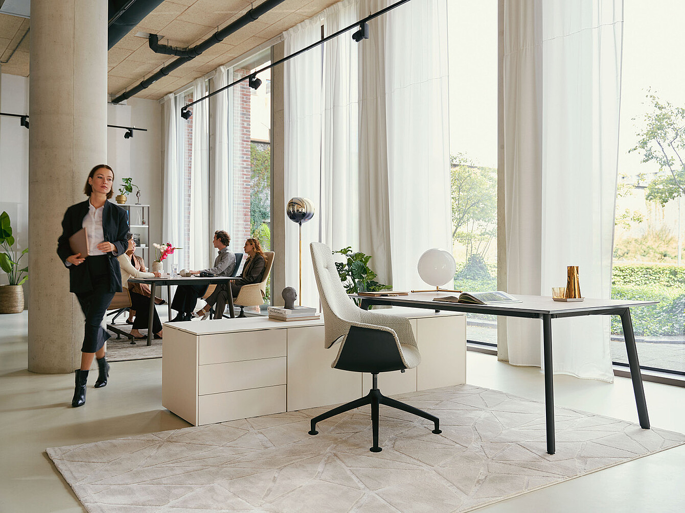 Modern office space with large windows, white curtains, a desk with a white Sedus se:mission swivel chair, and people sitting and walking around