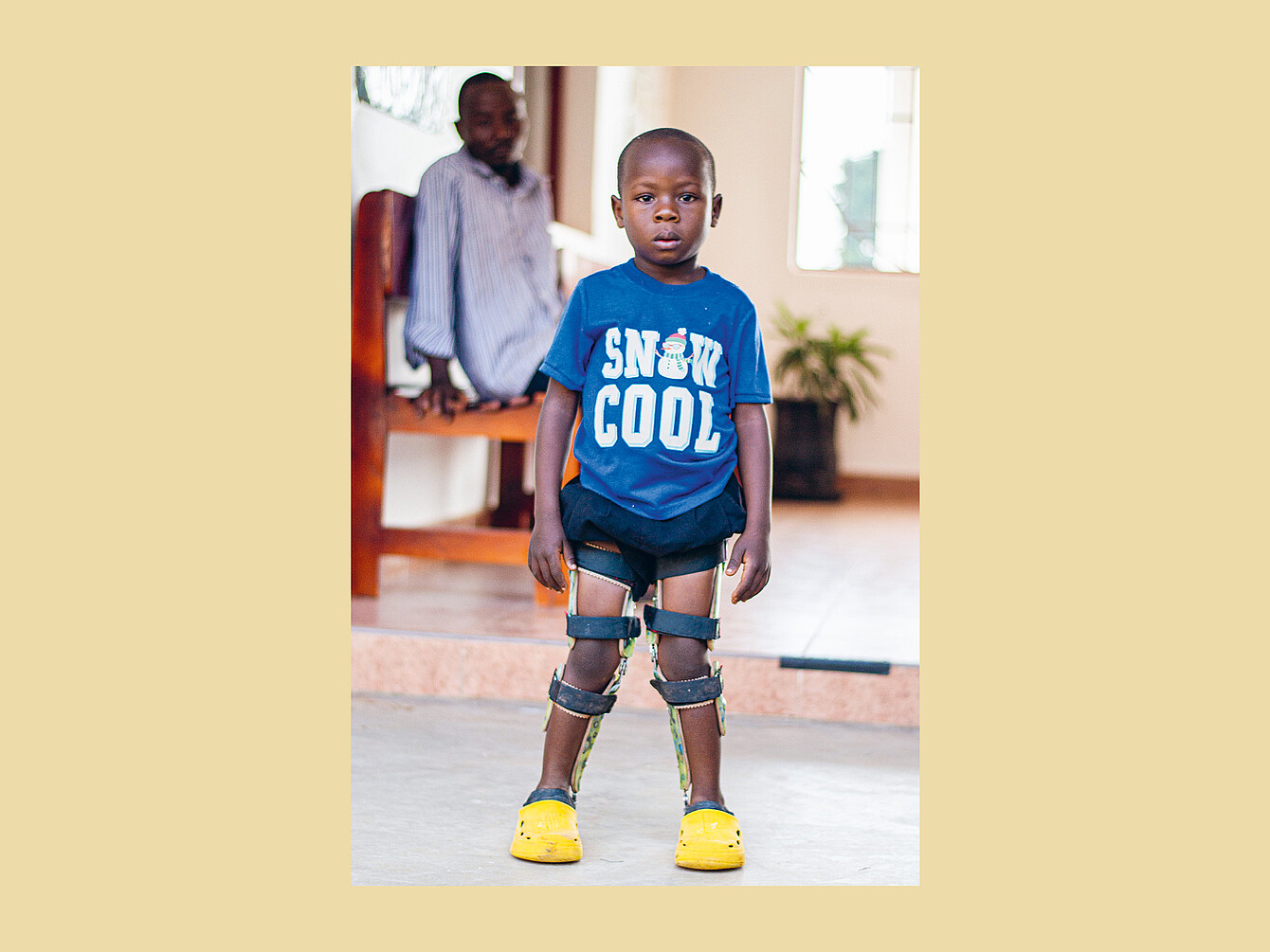 Kind steht mit Beinorthesen und gelben Schuhen in einem hellen Raum, trägt blaues T-Shirt mit weißer Aufschrift 'SNOW COOL'.