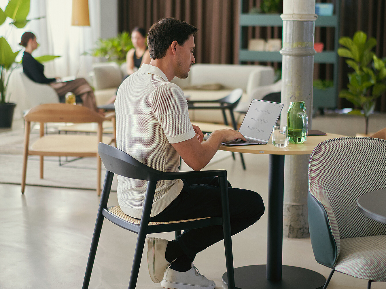 Person sitting at a round table in a work café, using a laptop with a glass of water and a green bottle nearby, illustrating varied atmospheres that boost employee engagement.