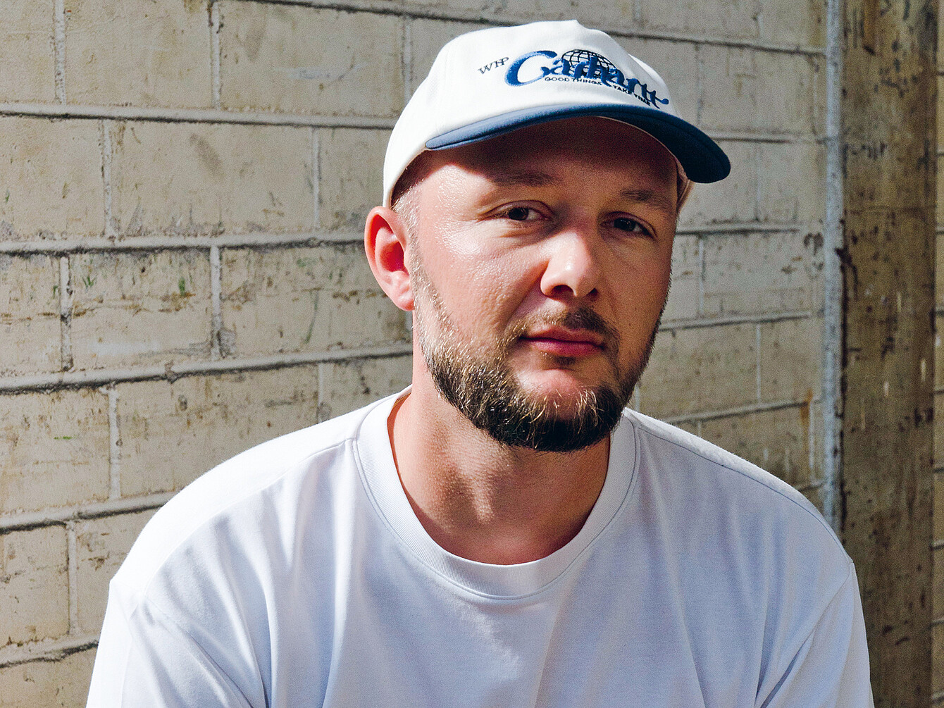 Man with a beard wearing a white cap and white t-shirt, sitting against a textured brick wall, representing designer Konstantin Thomas.