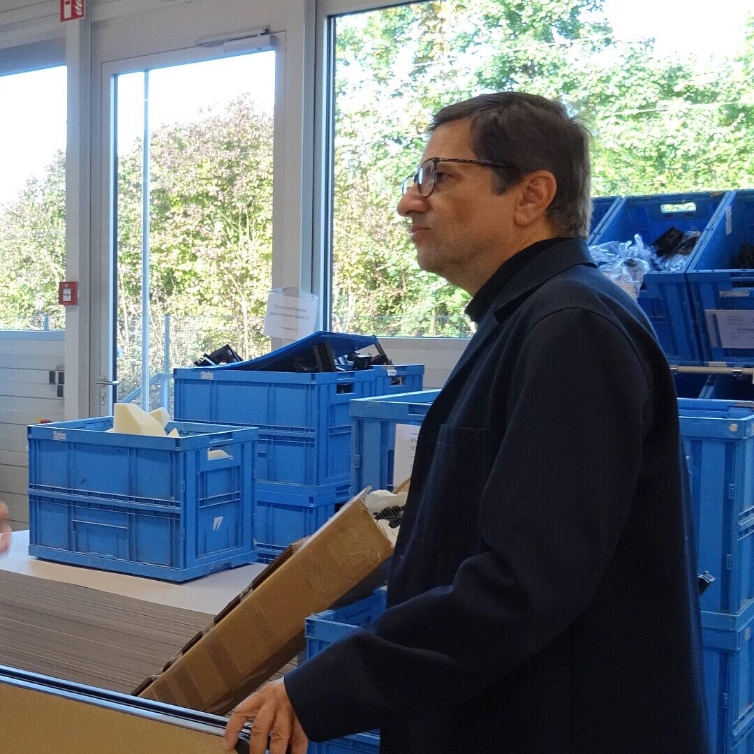 Zwei Männer (Konstantin Thomas und Rudi Raschke) stehen in einem Lager mit blauen Kunststoffboxen und sprechen miteinander, einer trägt eine Jeansjacke und eine Mütze, der andere einen dunklen Pullover.