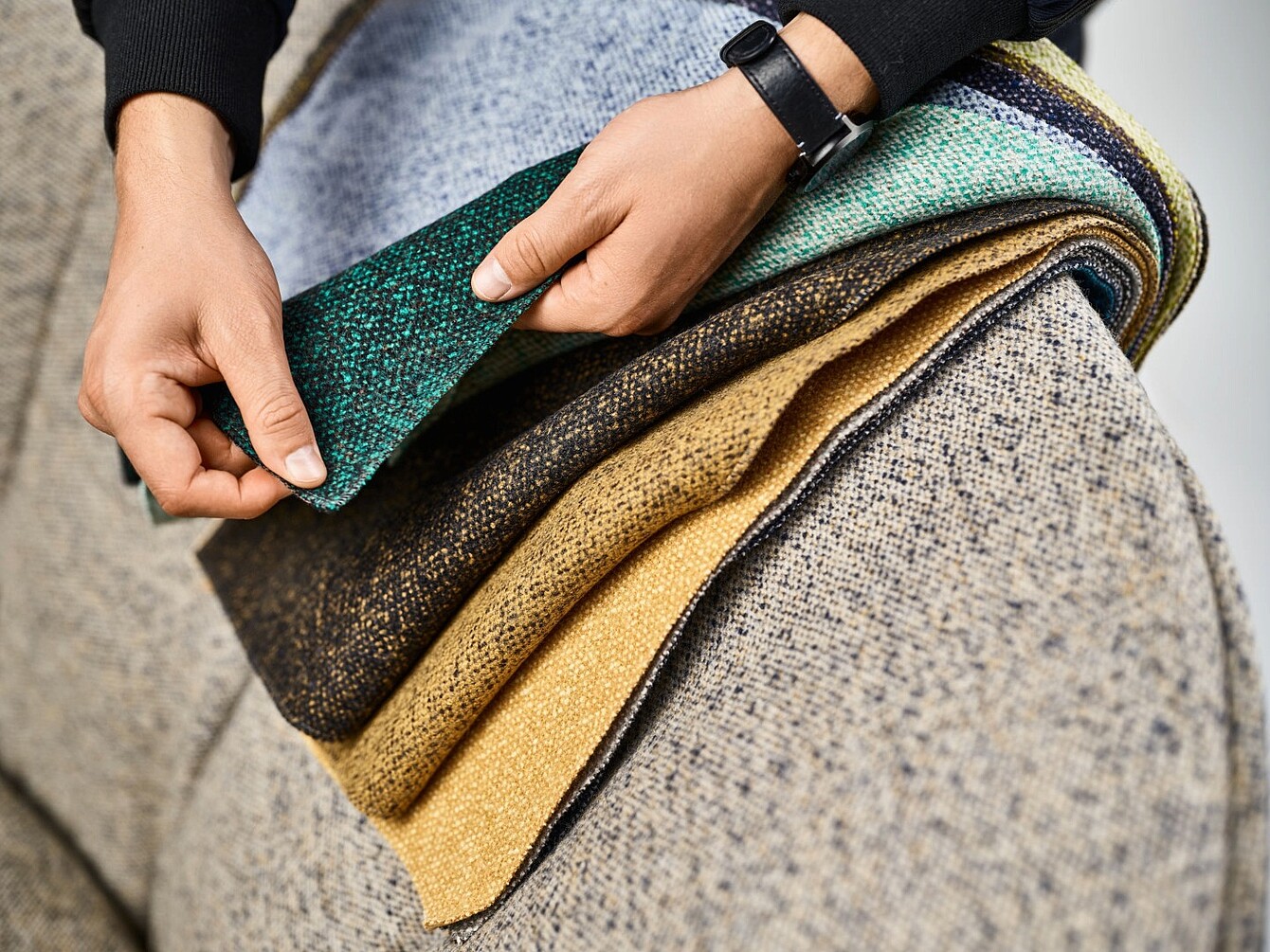 Hands holding and flipping through a stack of upholstery fabric samples in various textured patterns and colors