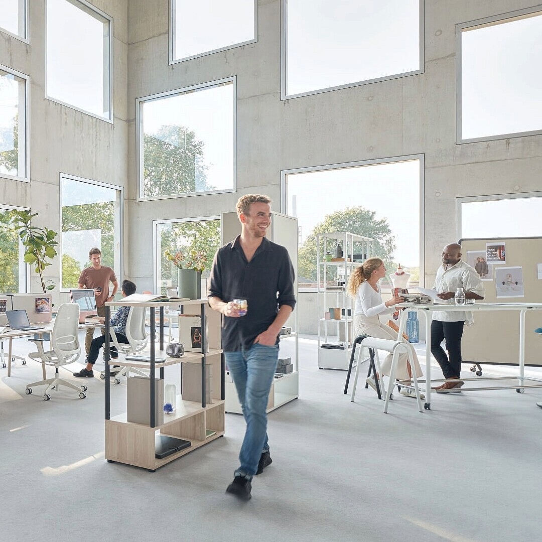 Helles, modernes Büro mit großen Fenstern, mehreren Mitarbeitenden an Tischen und Stehpulten, Pflanzen und offenem Raumkonzept