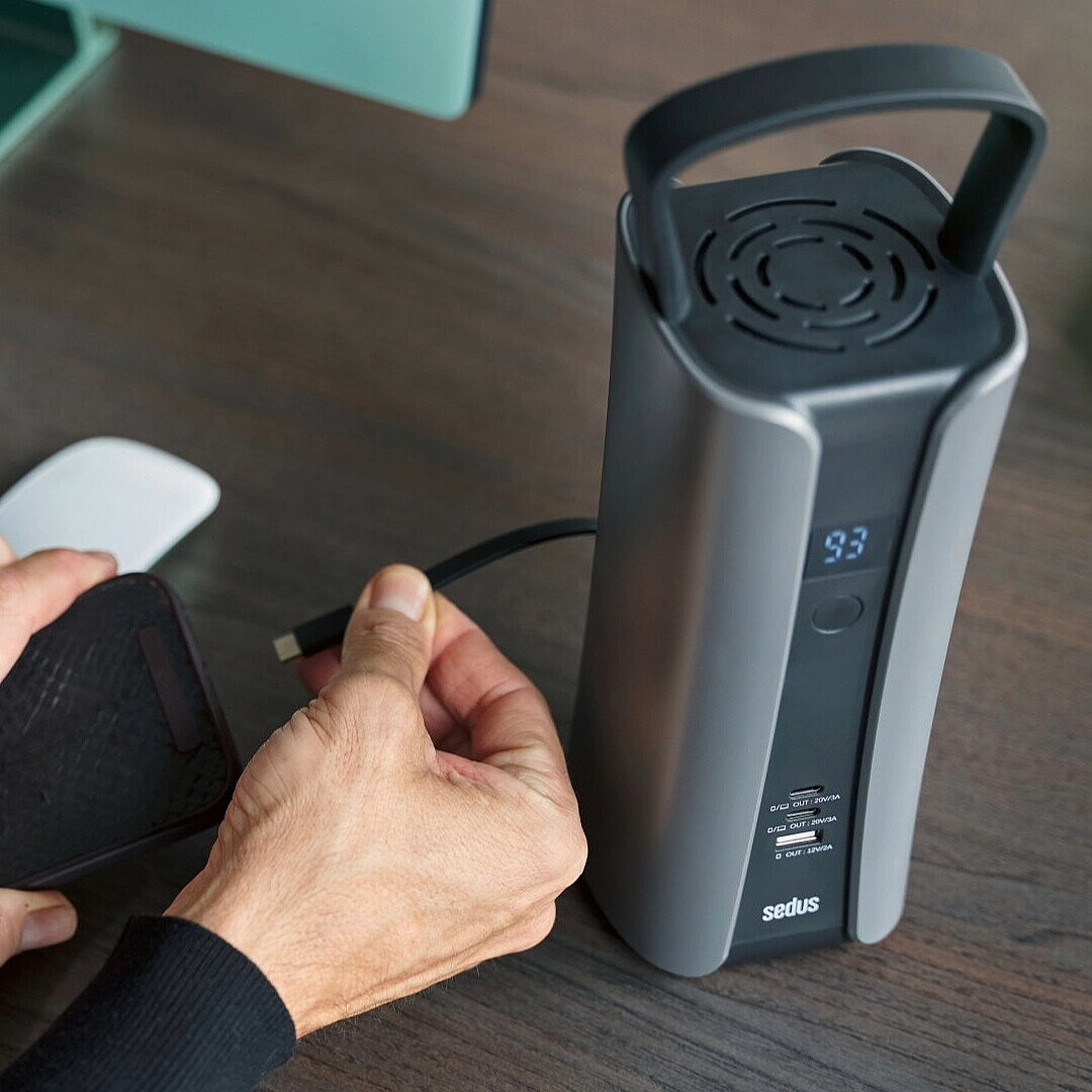 Hand hält USB-C-Kabel und steckt es in einen mobilen Akku mit Display auf einem Schreibtisch mit Tastatur, Maus und Smartphone.