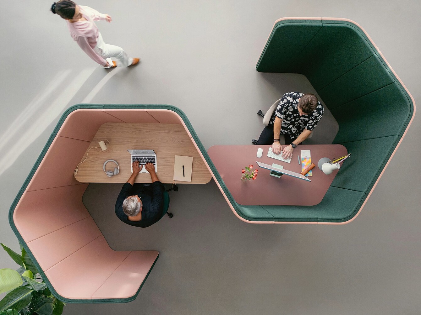 Zwei Personen sitzen an modernen, halbhohen Büroarbeitsplätzen mit gepolsterten Trennwänden in U-Form, eine weitere Person geht auf dem grauen Boden vorbei.