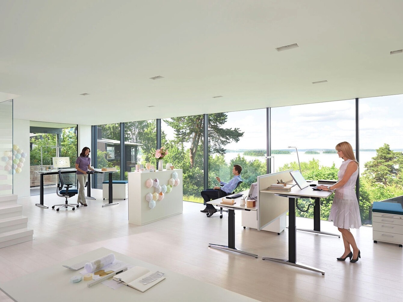 Helle, moderne Bürofläche mit großen Fenstern, in der drei Personen an höhenverstellbaren Schreibtischen arbeiten, zwei stehend und einer sitzend.