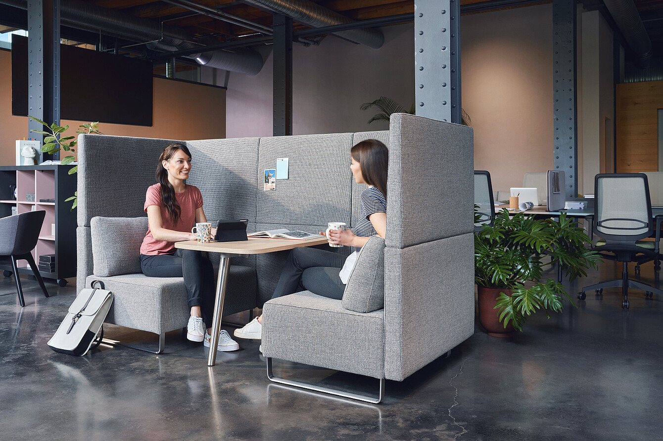 Two women sitting in a high-backed gray booth in an open office space talking and holding mugs