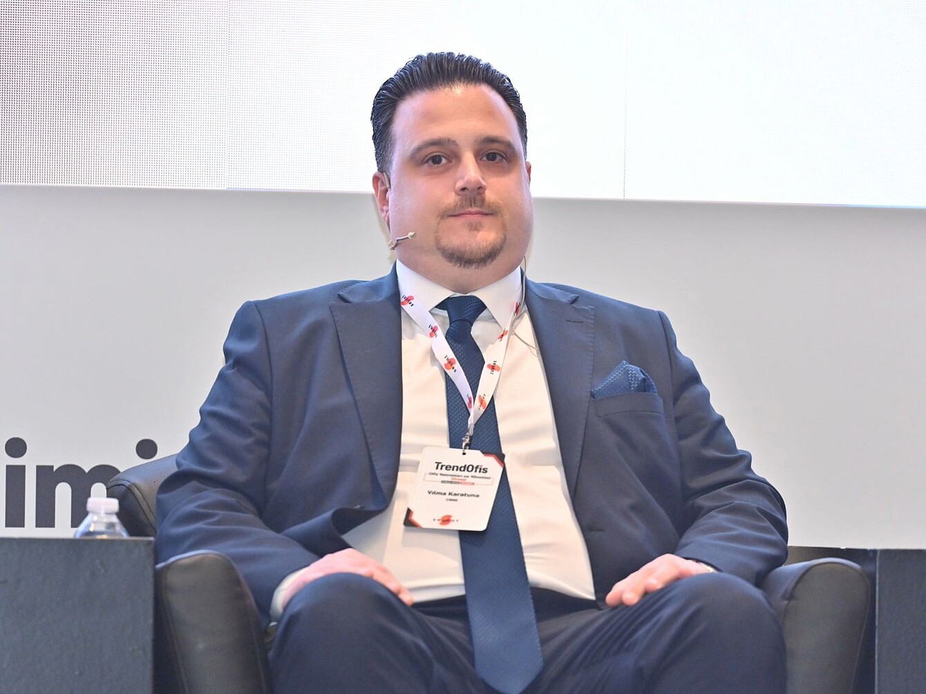 Mann in dunklem Anzug mit Krawatte sitzt auf schwarzem Sessel und trägt Namensschild mit Aufschrift 'TrendOfis'